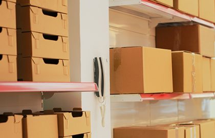 Empty warehouse with landline phone on wall, shelves and racks filled with merchandise in cardboard boxes. Multiple products prepared for shipment and transportation, stock goods.